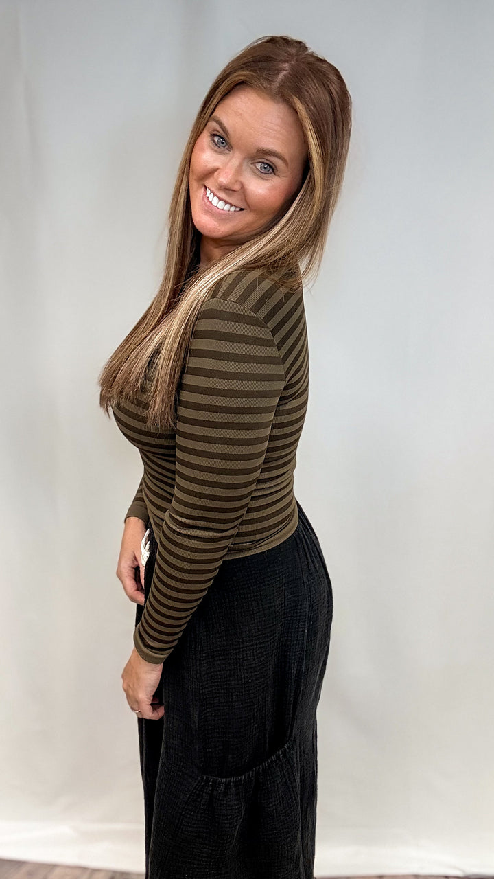 Woman wearing olive green stripe long sleeve top and black pants on plain background