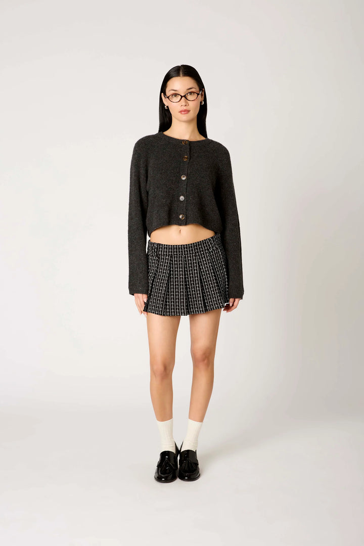 A model stands against a light gray studio background wearing a dark charcoal cropped button-front cardigan, a black-and-white micro-check pleated mini skirt, white socks, and black loafers.