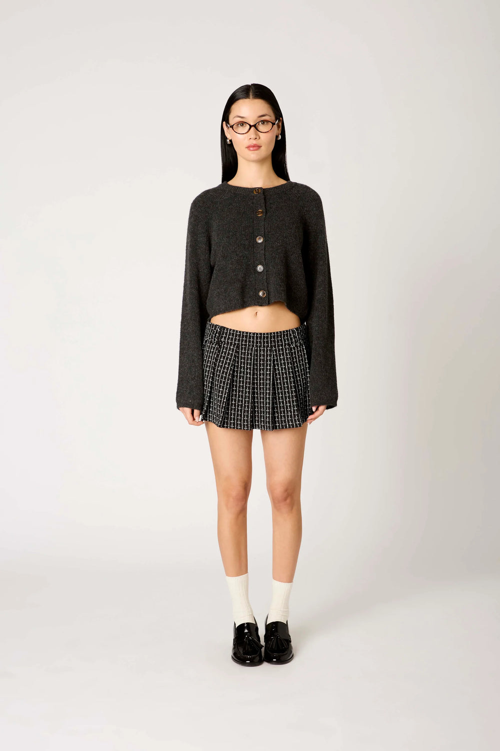 A model stands against a light gray studio background wearing a dark charcoal cropped button-front cardigan, a black-and-white checkered pleated mini skirt, white socks, and black loafers.
