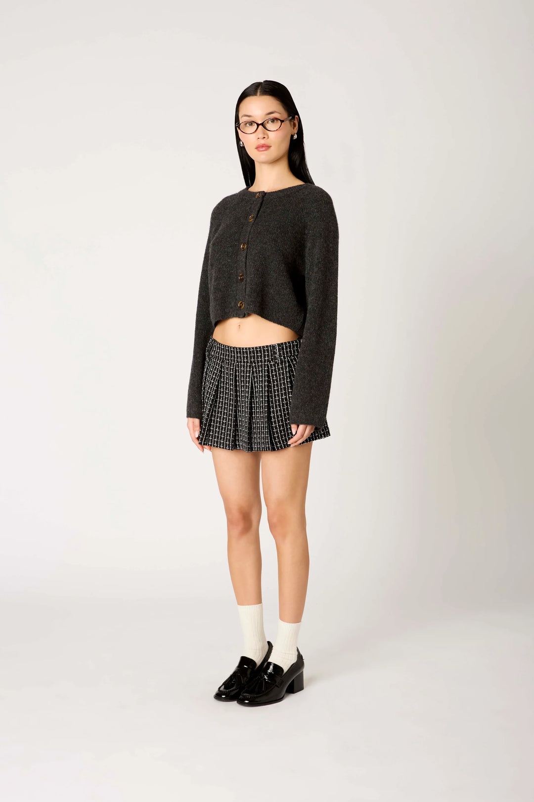 A model stands against a light gray studio background wearing a dark charcoal cropped cardigan, a black and white patterned pleated mini skirt, white socks, and black loafers.