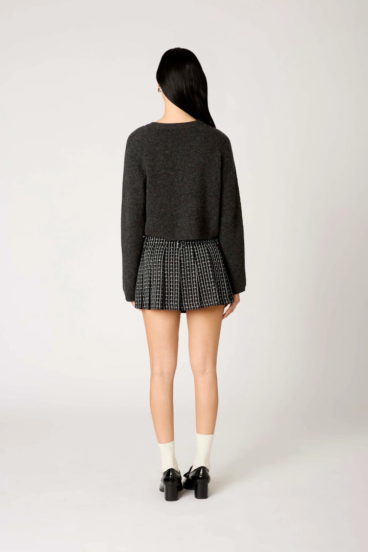 A model shown from the back wearing a dark gray knit sweater and a black and white houndstooth pleated mini skirt, paired with white socks and black shoes, against a light gray studio background.