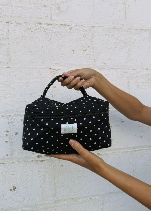 Woman holding black polka dot makeup bag in front of white brick wall