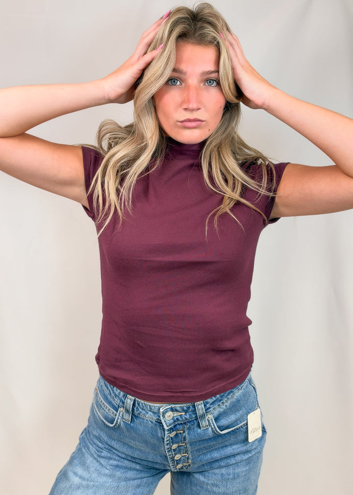 Woman wearing burgundy cap sleeve mock neck top with jeans on plain background