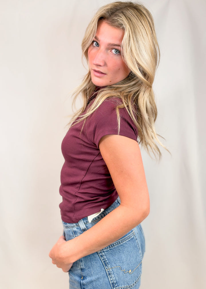 Woman shown from side view wearing burgundy cap sleeve mock neck top with jeans on plain background