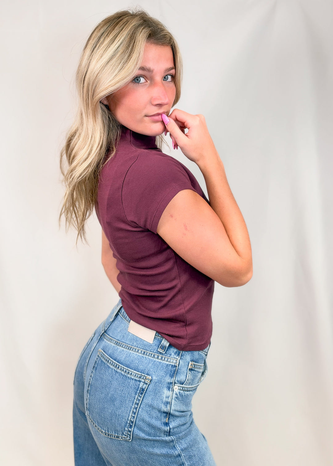 Woman shown from side view wearing burgundy cap sleeve mock neck top with jeans on plain background