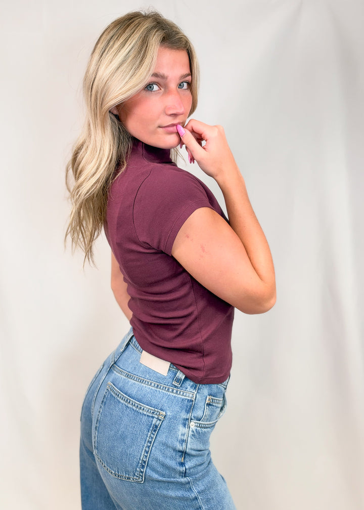 Woman shown from side view wearing burgundy cap sleeve mock neck top with jeans on plain background
