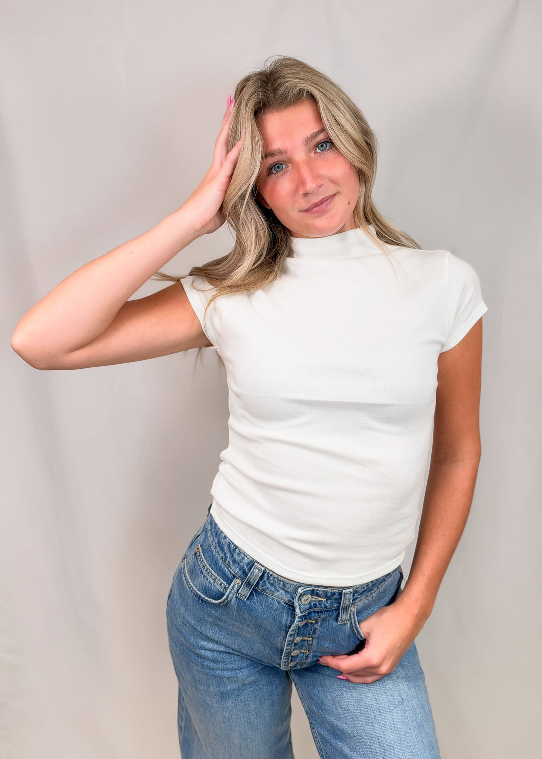 Woman wearing white cap sleeve mock neck top with jeans on plain background
