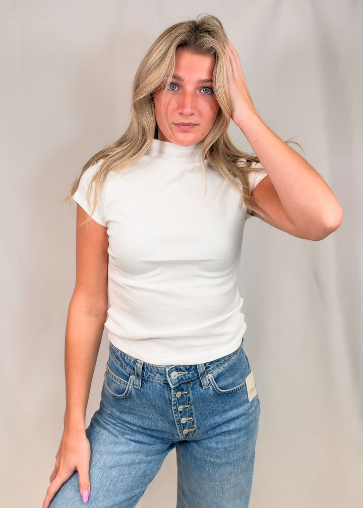Woman wearing white cap sleeve mock neck top with jeans on plain background