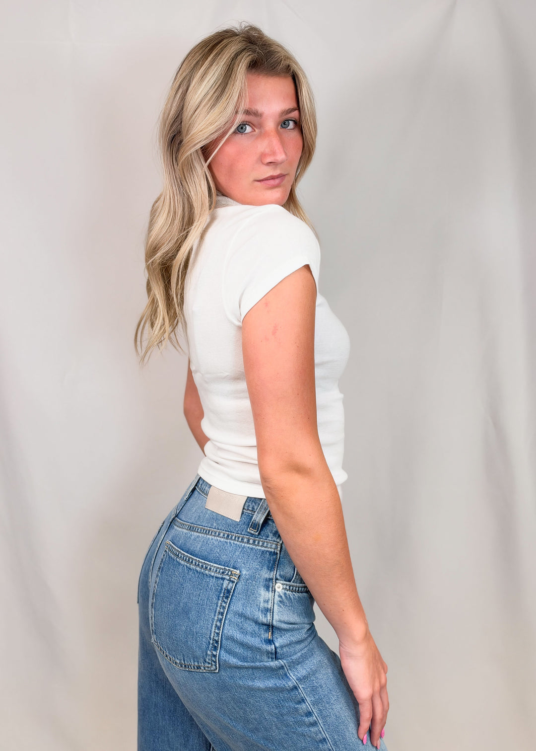 Woman from side view wearing white cap sleeve mock neck top with jeans on plain background