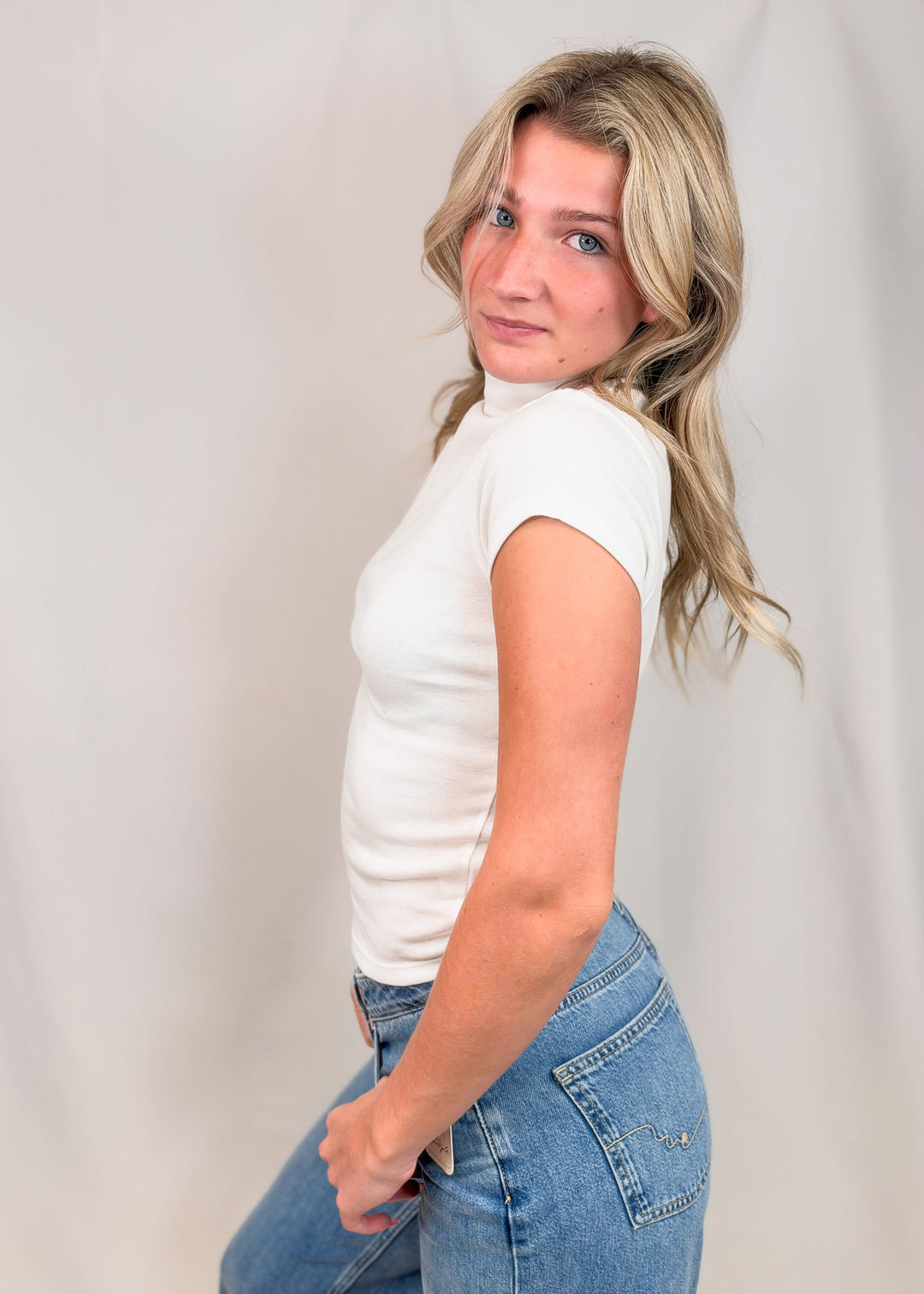 Woman shown from side view wearing white cap sleeve mock neck top with jeans on plain background