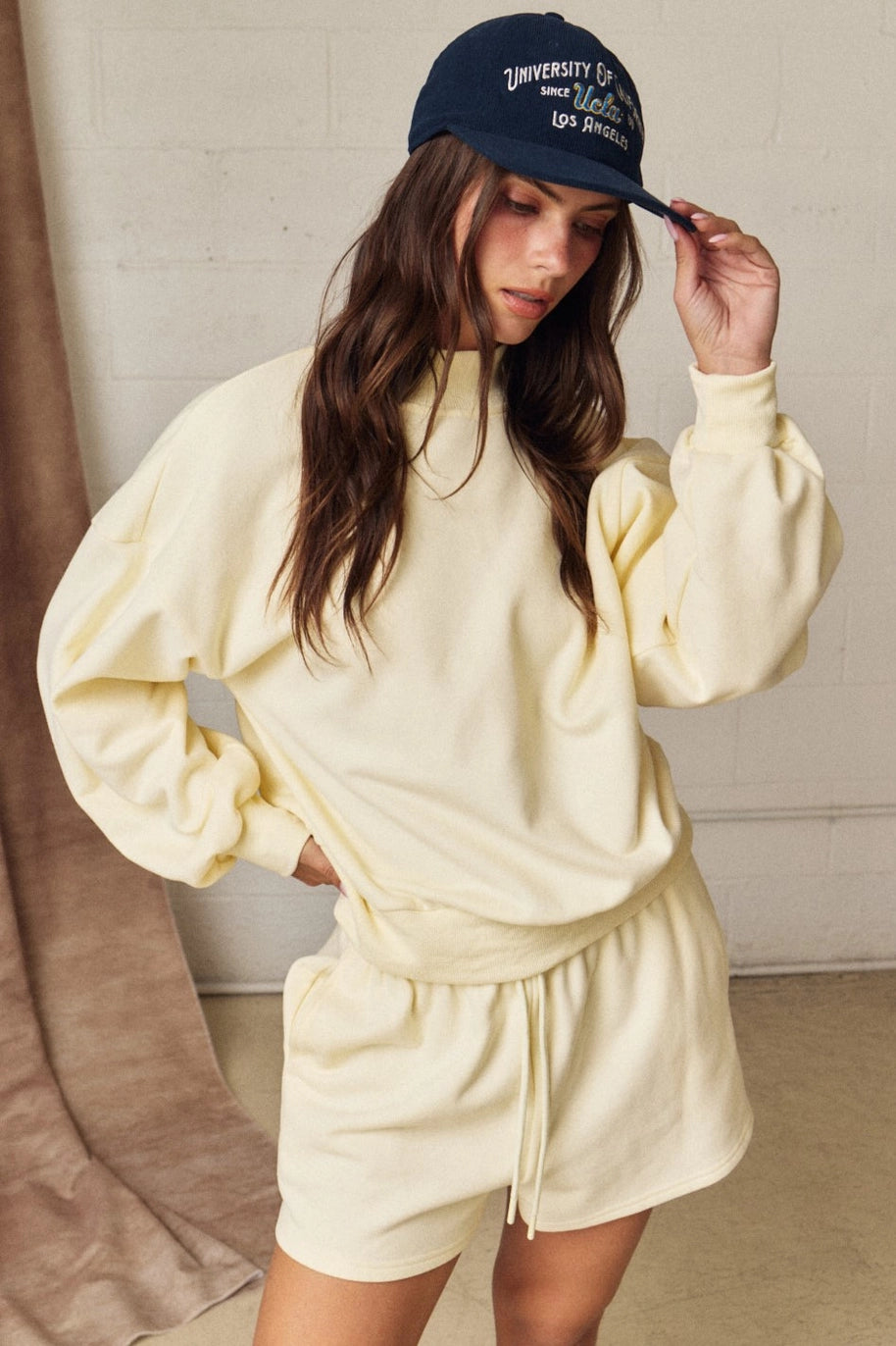 Woman wearing a yellow sweatshirt and shorts set with a navy cap indoors.