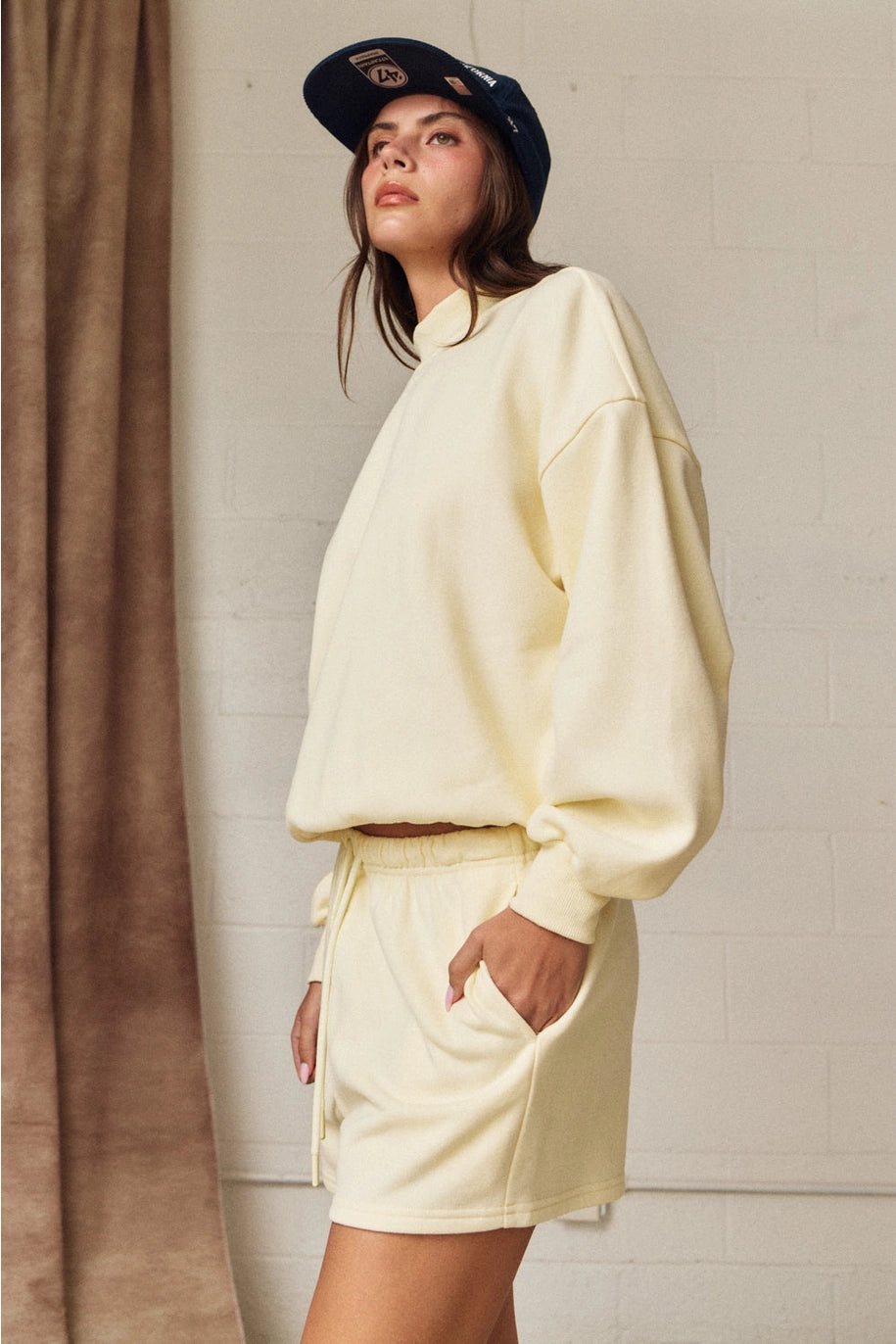 Woman wearing a light yellow sweatshirt and shorts set with a navy cap, standing against a neutral background.