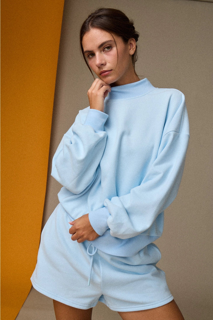 Woman wearing a light blue sweatshirt and shorts set against a gradient wall.