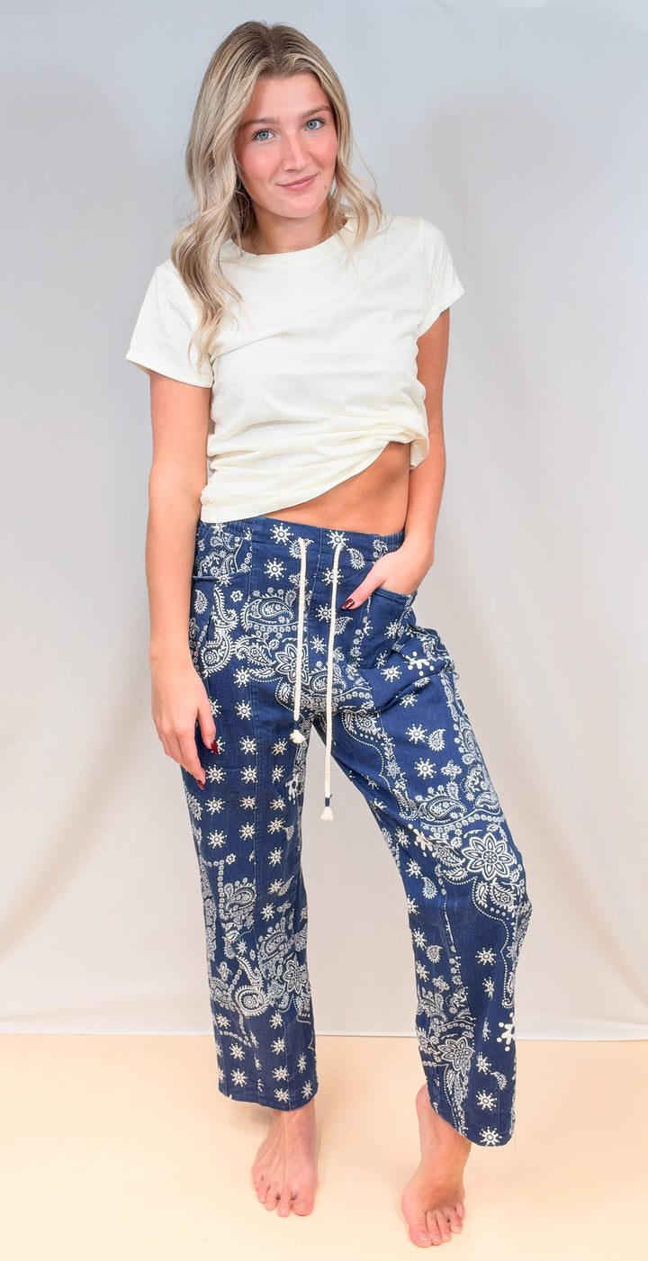 Woman wearing cream tee with blue paisley pull on pants on plain background