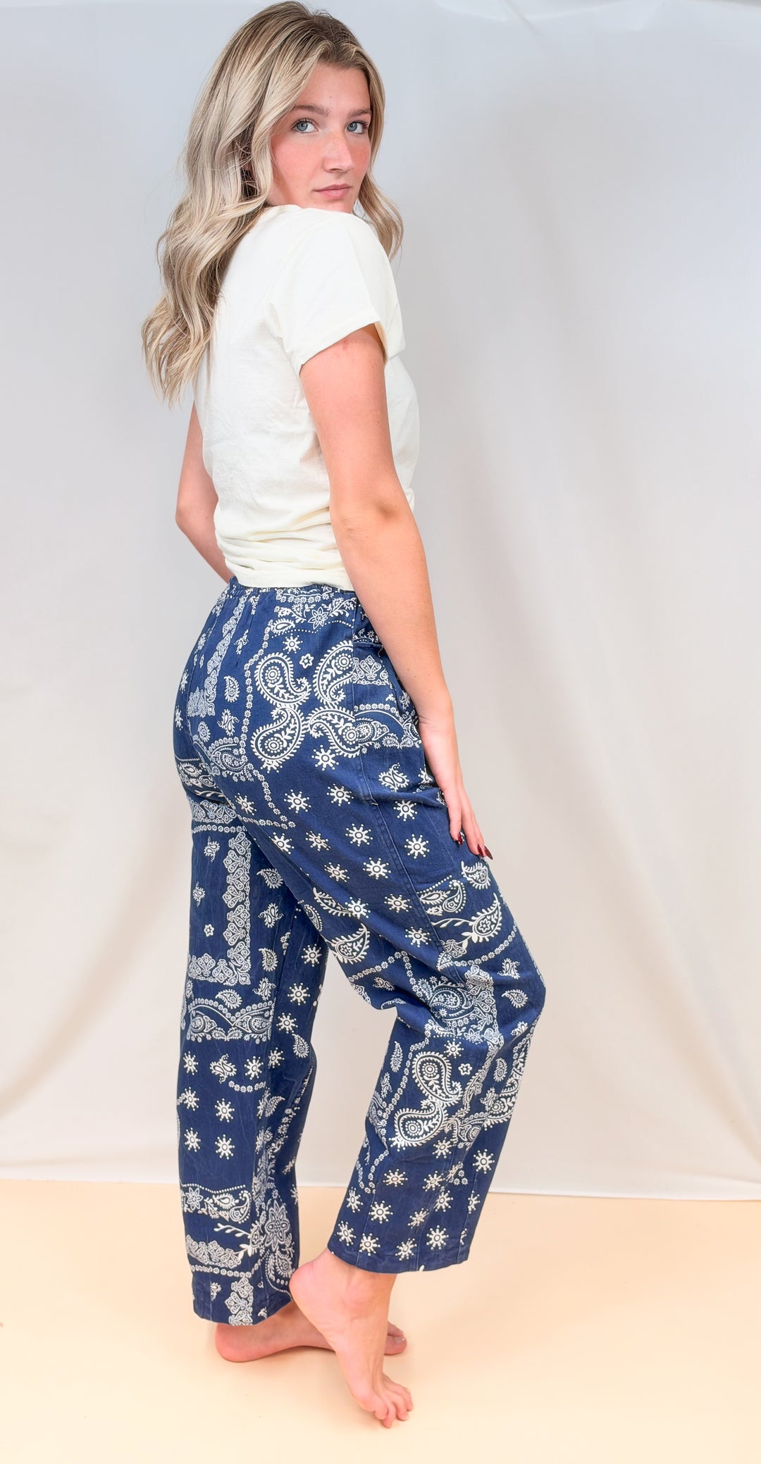 Woman shown from side view wearing cream tee with blue paisley pull on pants on plain background