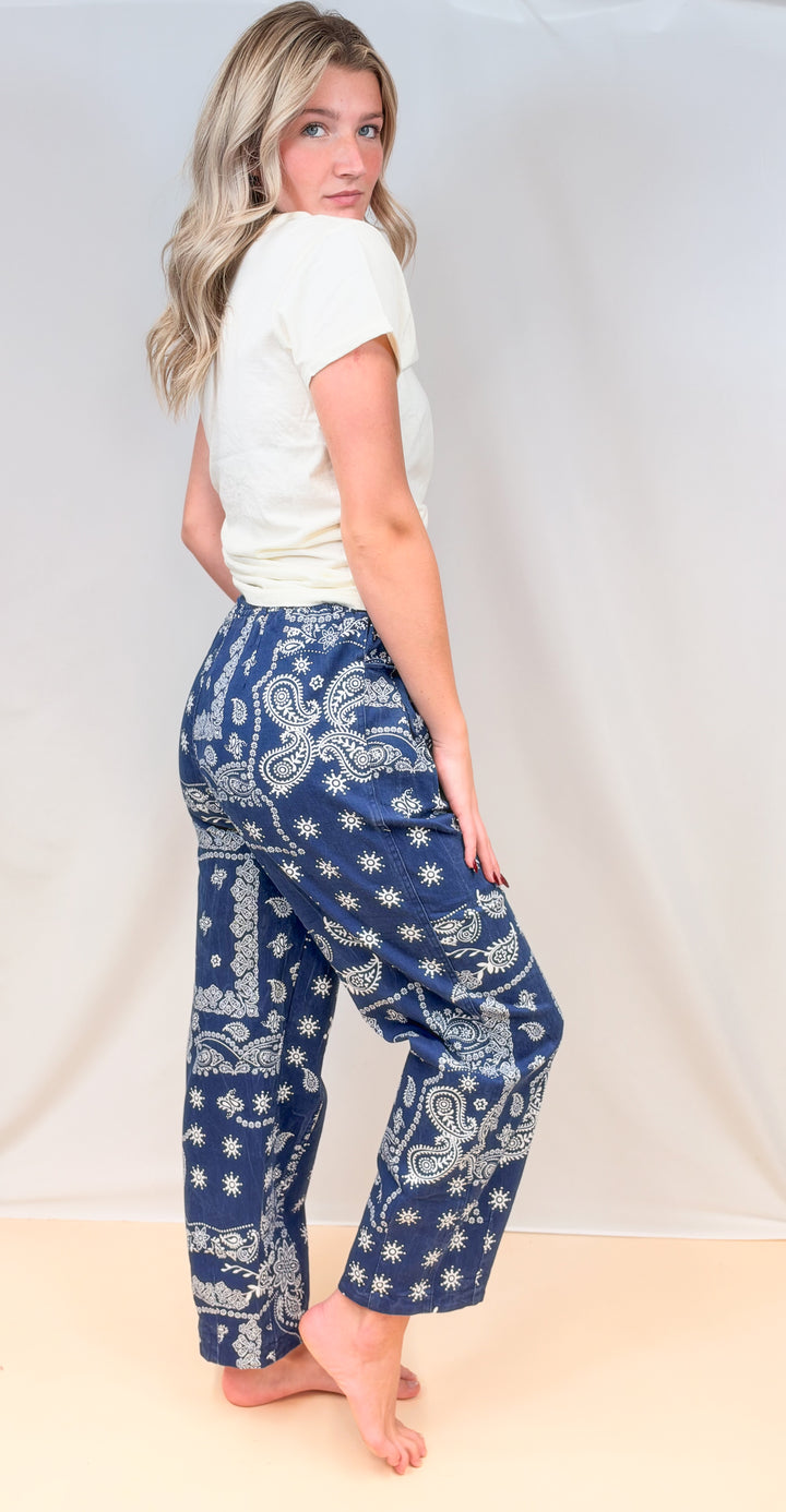 Woman shown from side view wearing cream tee with blue paisley pull on pants on plain background