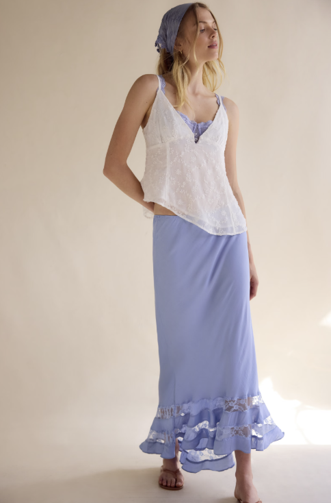 Woman wearing a white top and light blue skirt with a headscarf on a plain background