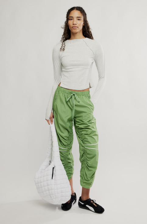 Woman wearing a white long-sleeve top and green pants, holding a white bag on a light gray background
