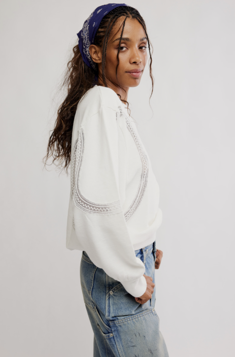 A woman wearing a white cropped sweatshirt with silver chain embroidery on the sleeves, paired with light-wash blue jeans and a purple bandana, photographed against a plain light gray background.