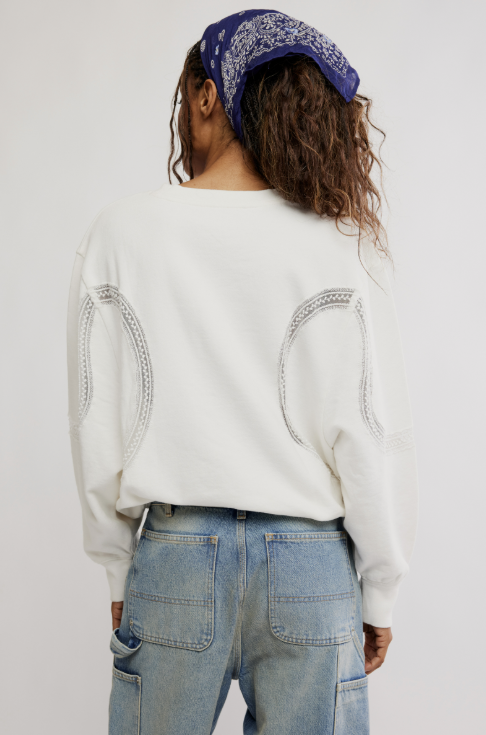 A person shown from the back wearing a white long-sleeve sweatshirt with decorative silver circular embroidery on the shoulders, paired with light blue jeans and a blue patterned bandana.