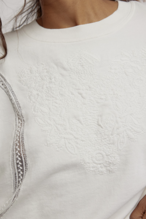Close-up of a white crewneck sweatshirt featuring a tonal floral jacquard pattern on the front and a sheer embroidered lace detail along the side.