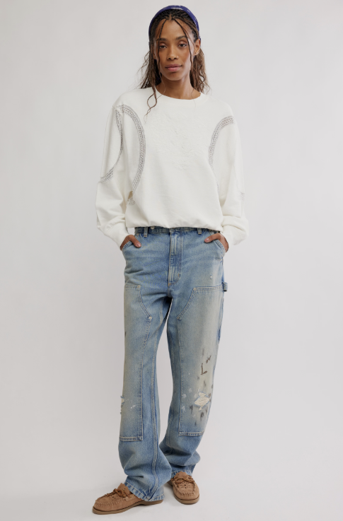 A model stands against a light gray studio background wearing a white crewneck sweatshirt with textured details, light-wash blue distressed jeans, and brown suede shoes.