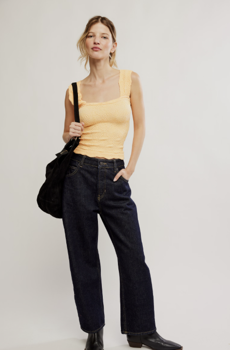 Woman wearing a yellow tank top and black jeans holding a black bag on a plain background