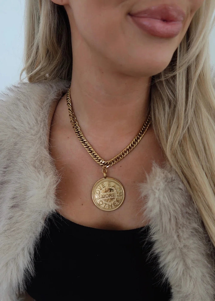 Person wearing a gold necklace with a large pendant, dressed in a black top and light fur coat.