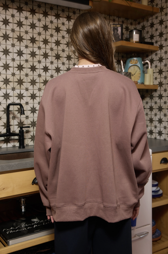 A person viewed from behind wearing an oversized dusty rose crewneck sweatshirt in a kitchen with star-patterned tiles and wooden shelves.