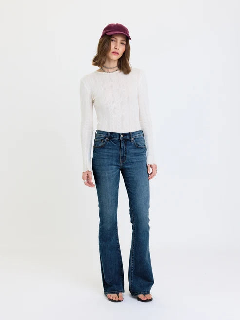 A model stands against a plain white studio background wearing a cream-colored long-sleeve knit top, dark blue flare jeans, a burgundy cap, and black sandals.