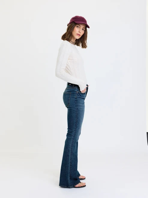 A woman modeling a pair of dark blue flared jeans with a white long-sleeve top and a burgundy baseball cap, standing against a plain white studio background.