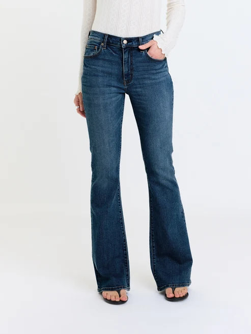 A person wearing dark blue flared denim jeans with a white ribbed long-sleeve top and black sandals, shown from the waist down against a plain white studio background.