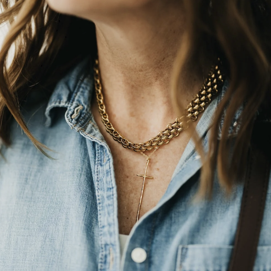 A close-up lifestyle shot of a person wearing a layered gold necklace set, featuring a textured chain and a delicate cross pendant, styled with a light blue denim shirt.