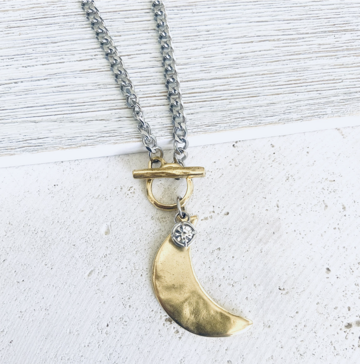 A silver chain necklace with a gold-toned toggle clasp and a crescent moon pendant, accented by a small clear gemstone, displayed on a textured white surface.