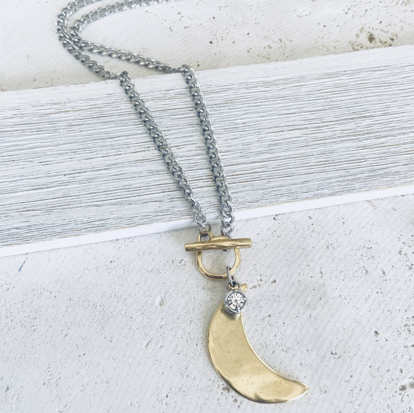 A silver chain necklace with a toggle clasp and a gold crescent moon pendant, accented with a small clear stone, displayed on a textured white surface.