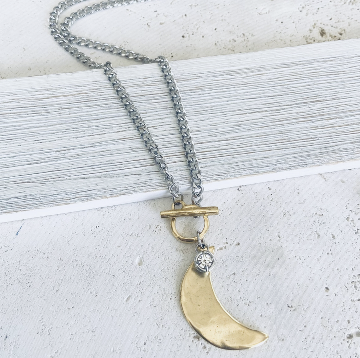 A silver chain necklace with a toggle clasp and a gold crescent moon pendant, accented with a small clear stone, displayed on a textured white surface.