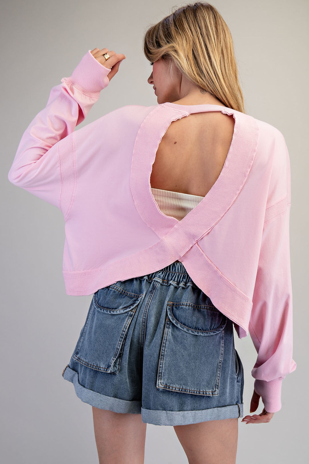 Person wearing a pink sweater with open back and denim shorts on a gray background