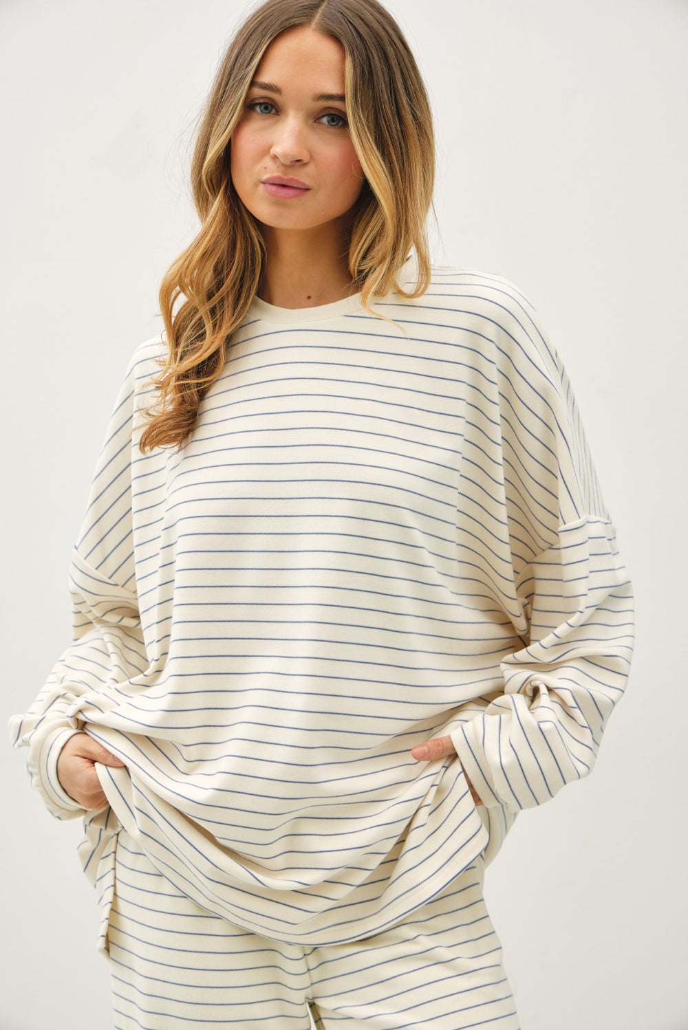 A woman wearing a cream-colored long-sleeve top with thin navy horizontal stripes, paired with matching striped pants, against a plain light gray background.