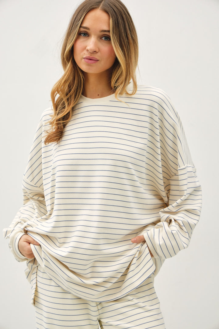 A woman wearing a cream-colored long-sleeve top with thin navy horizontal stripes, paired with matching striped pants, against a plain light gray background.