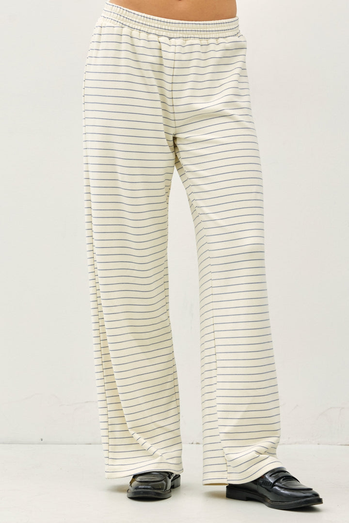 A person wearing cream-colored wide-leg pants with thin dark horizontal stripes, paired with black loafers, shown from the waist down against a plain off-white studio background.