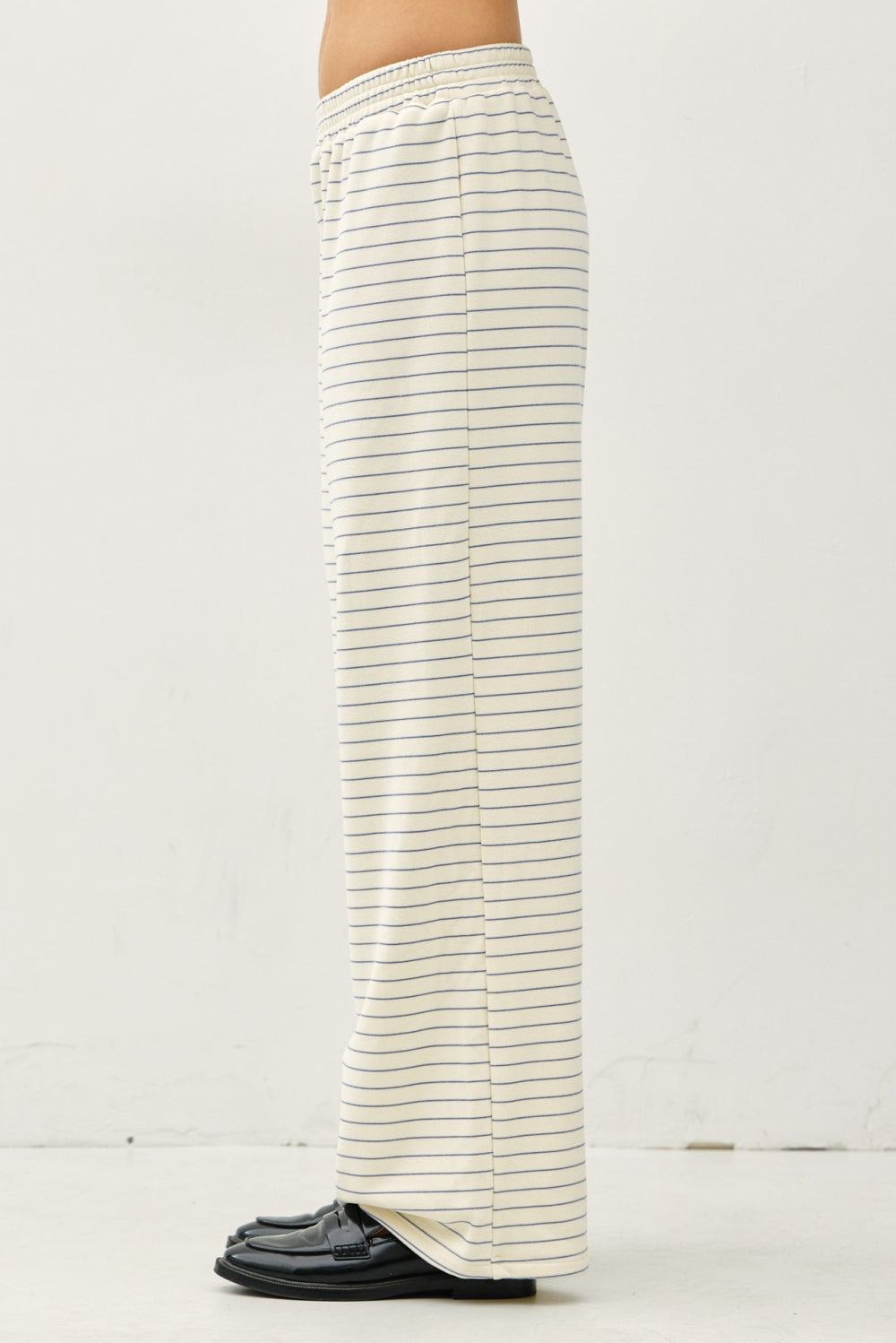A side view of a person wearing cream-colored wide-leg pants with thin green horizontal stripes, paired with black loafers, against a plain off-white wall.
