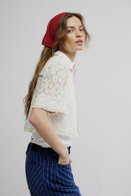 Woman wearing a white lace top and blue pants with a red headscarf on a plain background