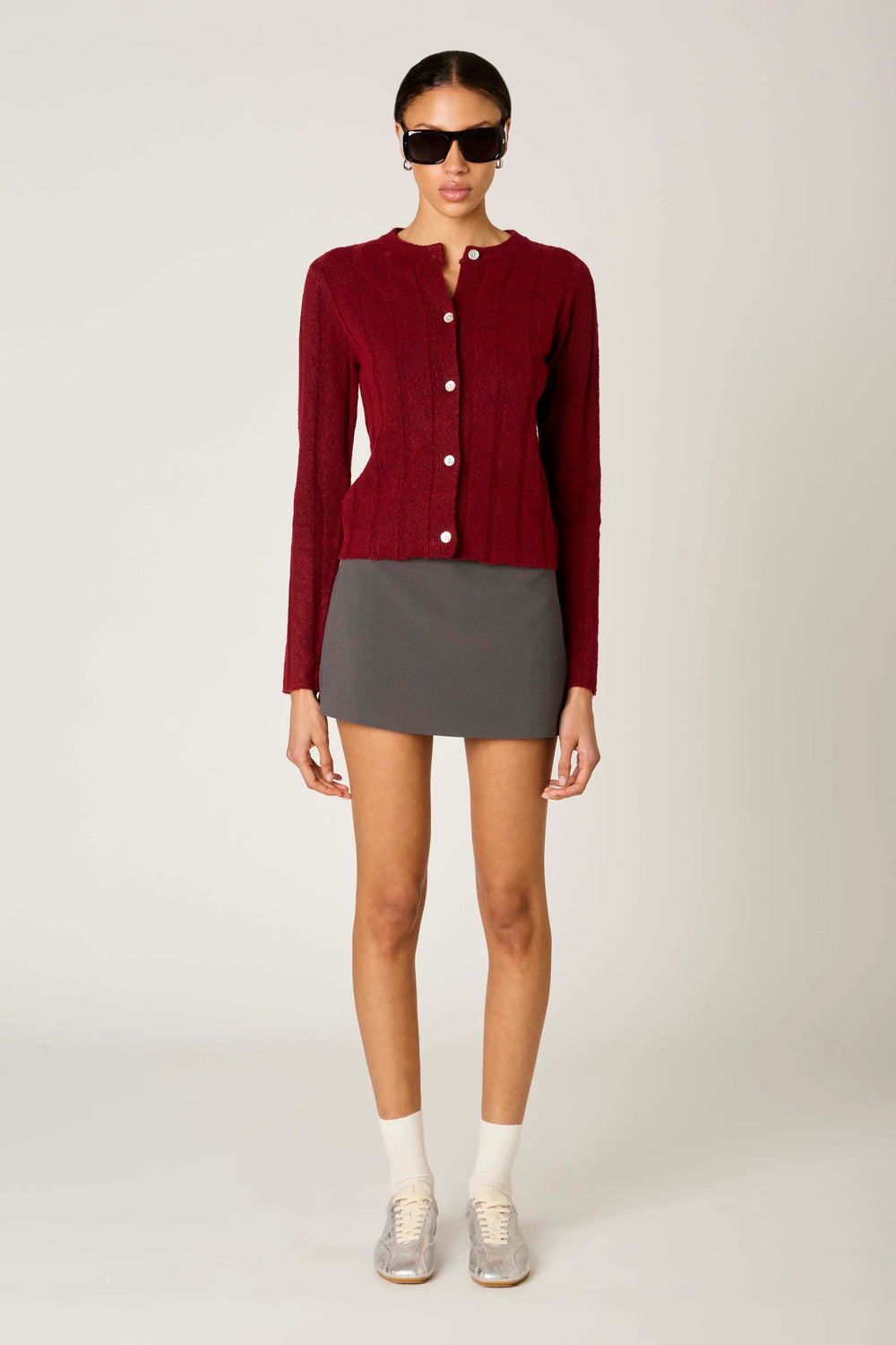 A model stands against a light gray studio background wearing a burgundy ribbed knit cardigan with white buttons, a dark gray mini skirt, white socks, and silver sneakers.
