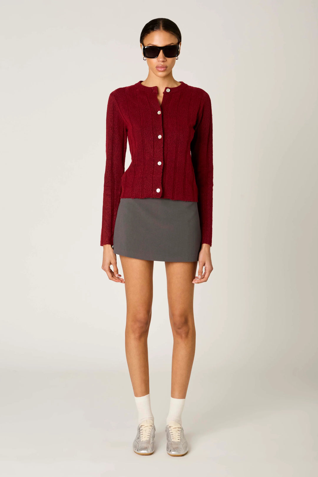 A model stands against a light gray studio background wearing a burgundy ribbed knit cardigan with white buttons, a dark gray mini skirt, white socks, and silver sneakers.