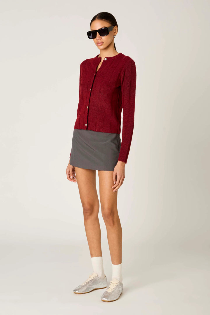 A model wearing a burgundy ribbed knit cardigan, a gray mini skirt, white socks, and light gray sneakers, standing against a light gray studio background.