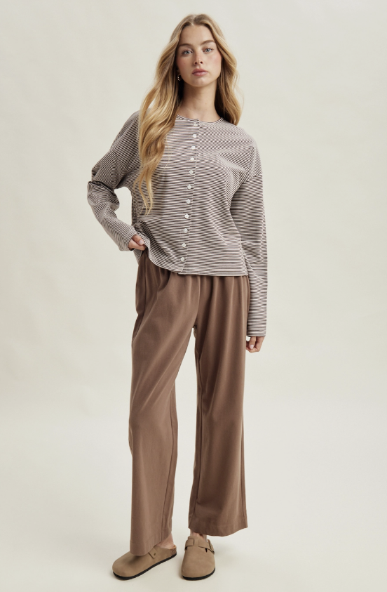 A woman modeling a two-piece lounge set consisting of a brown and white striped long-sleeve button-up top and brown wide-leg pants, paired with brown slip-on shoes, against a light beige studio background.