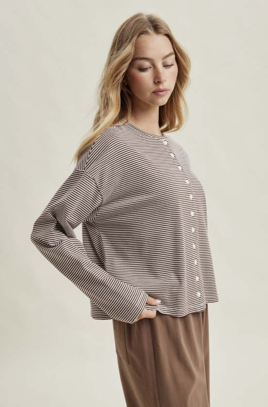 A woman wearing a long-sleeve button-front top with thin horizontal brown and white stripes, paired with brown pants, photographed against a plain light beige studio background.