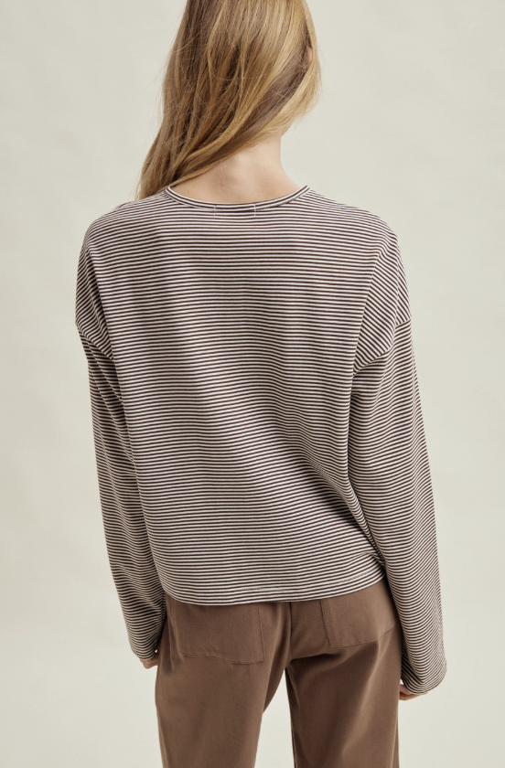 A woman shown from the back wearing a long-sleeve crewneck top with thin horizontal dark brown and beige stripes, paired with brown pants, against a light beige studio background.
