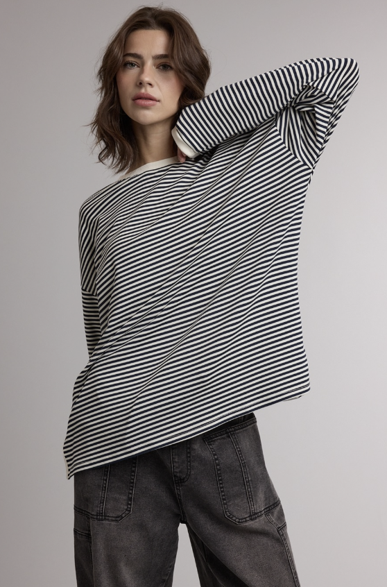 A woman modeling a black and white horizontally striped long-sleeve top with a relaxed, oversized fit, paired with dark gray jeans against a neutral gray studio background.