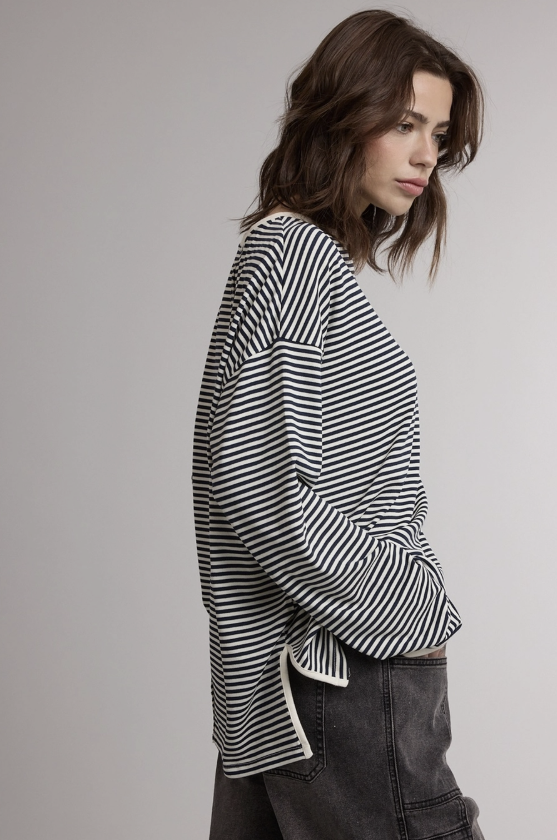 A woman wearing a black and white horizontally striped long-sleeve top with a side tie detail, paired with dark gray jeans, photographed against a neutral light gray background.